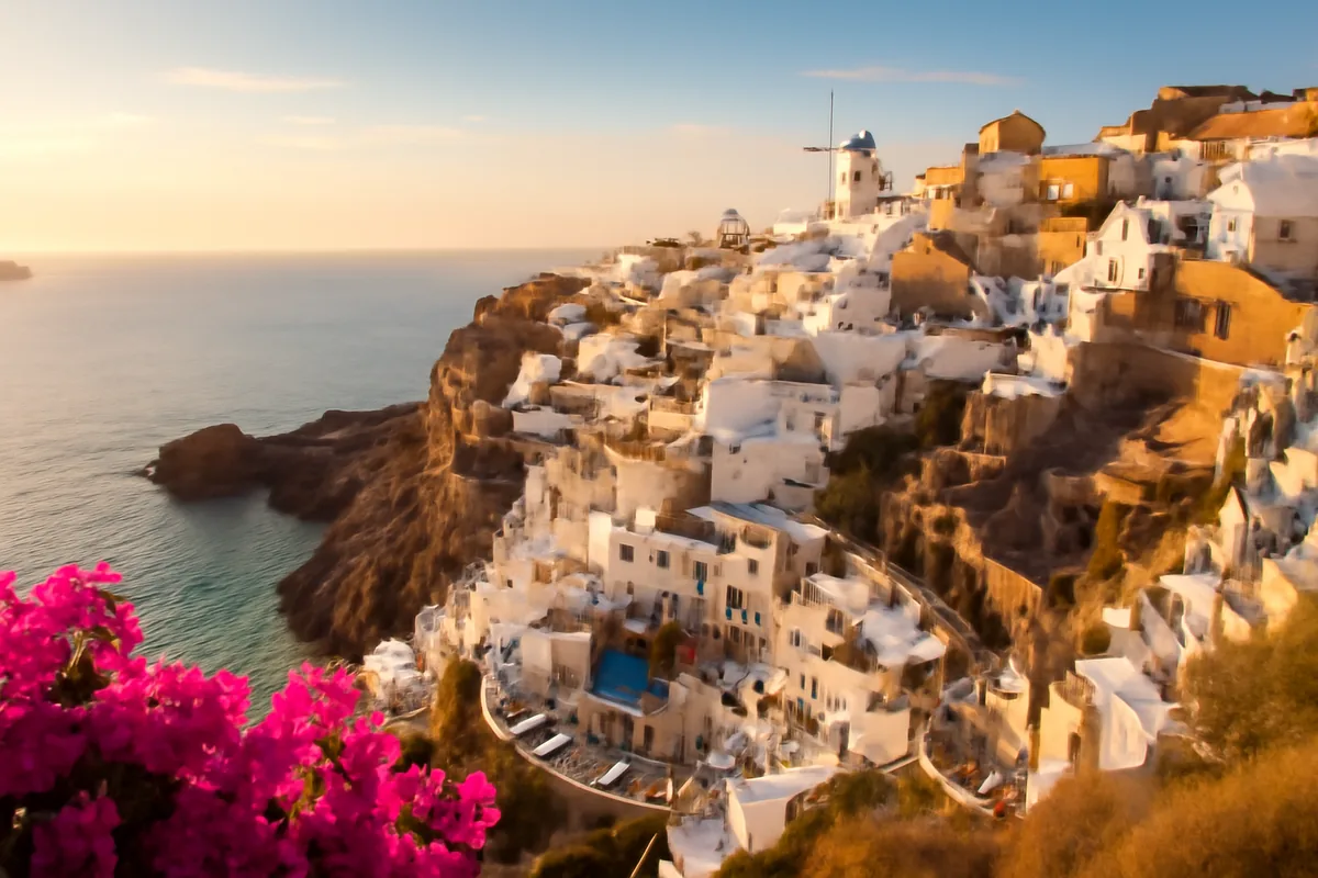 A breathtaking Mediterranean coastal view featuring whitewashed buildings on a clifftop, turquoise waters, and colorful bougainvillea flowers bathed in warm golden light